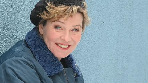 Eine Frau mit schwarzer Kappe, unter der blonde Locken hervorblitzen, sitzt vor einer blaugrauen Wand in der Hocke und lächelt in die Kamera.