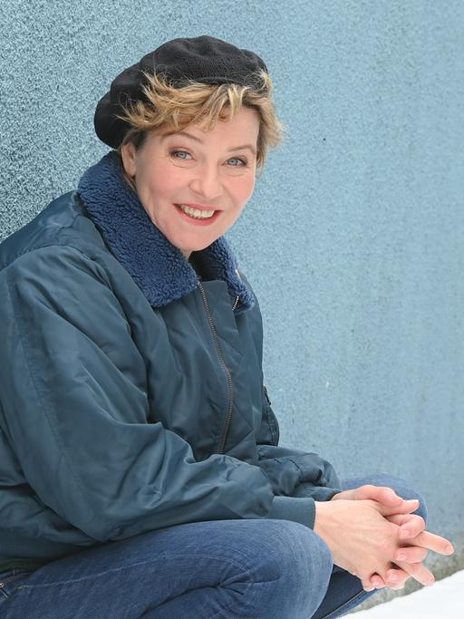 Eine Frau mit schwarzer Kappe, unter der blonde Locken hervorblitzen, sitzt vor einer blaugrauen Wand in der Hocke und lächelt in die Kamera.