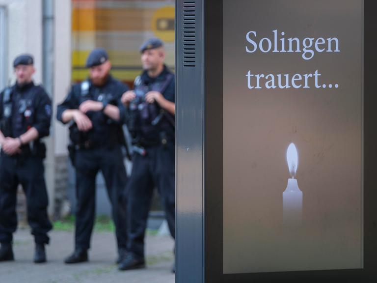 Polizisten stehen hinter einer Anzeigentafel "Solingen trauert..." mit Kerze im Gedenken an die Opfer des Attentats von Solingen am Tatort Fronhof am 01.09.2024.