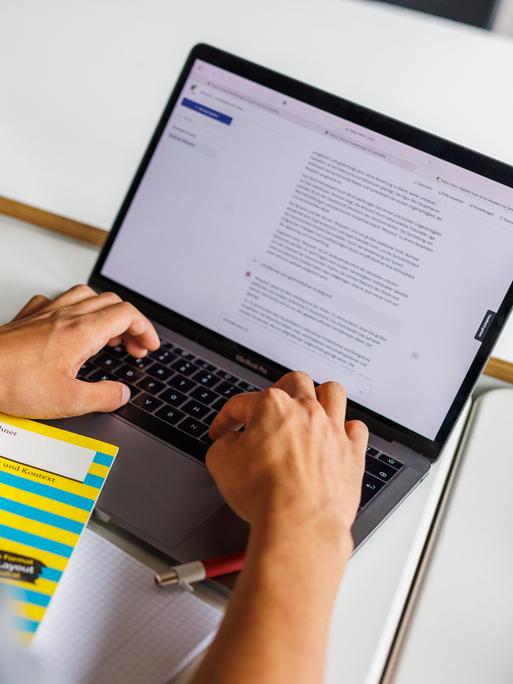 Ein Schüler sitzt vor einem Laptop und benutzt Chat-GPT. Vor ihm liegt ein gelbes Buch: "Woyzeck" von Büchner.
