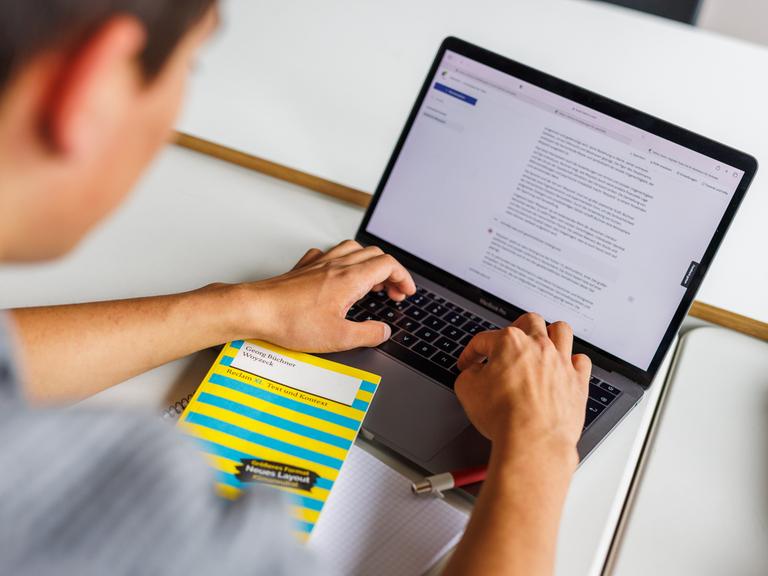 Ein Schüler sitzt vor einem Laptop und benutzt Chat-GPT. Vor ihm liegt ein gelbes Buch: "Woyzeck" von Büchner.