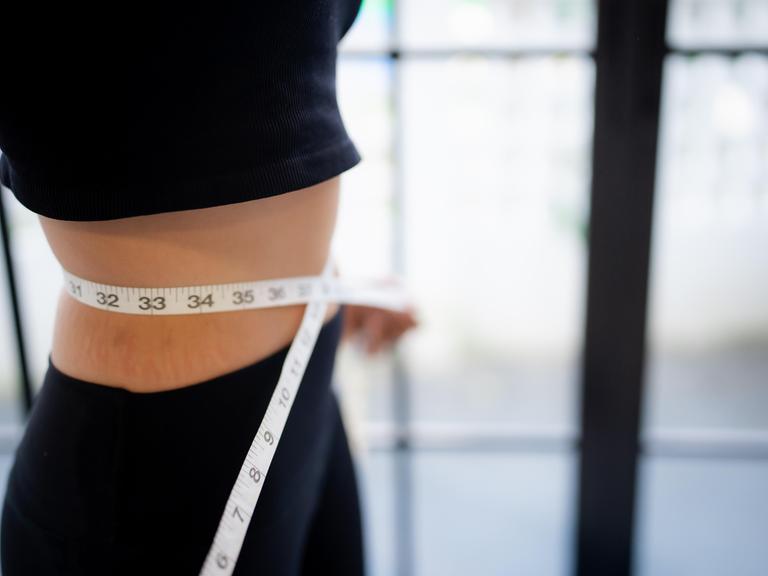 Eine schlanke Frau in Sportklamotten hält sich ein Maßband an die Taille.