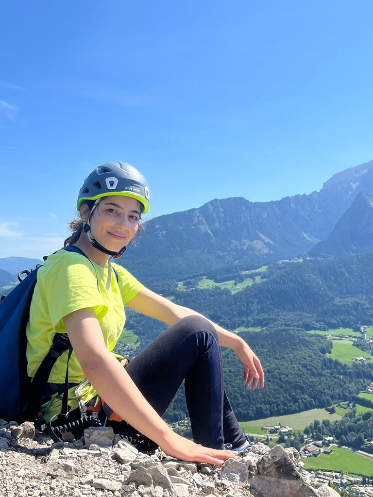 Eine Frau mit Helm und Rucksack sitzt auf einem Felsen, im Hintergrund ist ein Bergmassiv zu sehen.