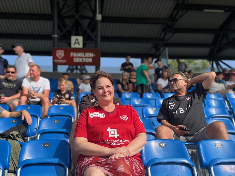 Frau mit roten Kleid sitzt in einem Fußballstadion mit blauen Sitzen, unter weiteren Zuschauern.