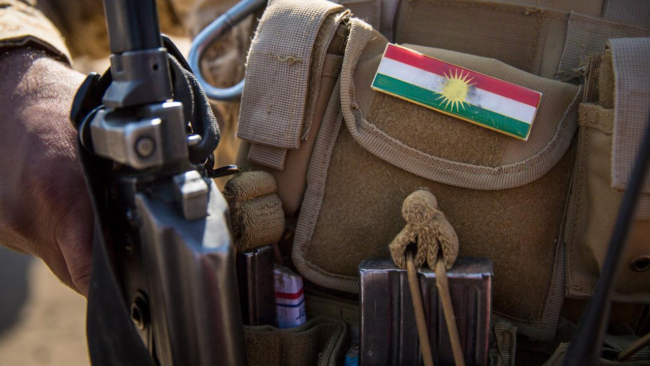 Die Nationalflagge von Kurdistan auf einer Uniform. Links daneben: eine Hand an einem Gewehr.