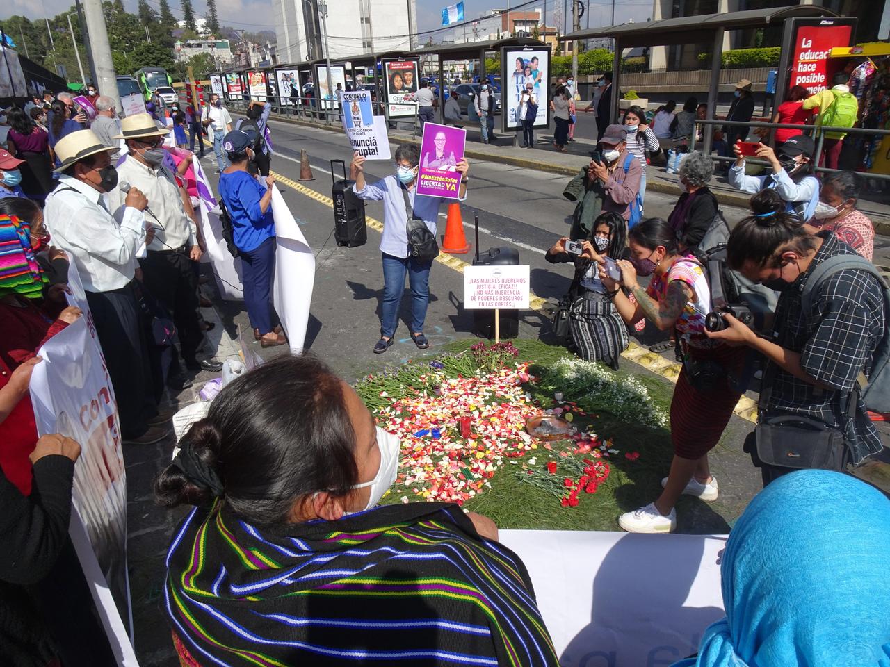 Straßenszene: Menschen mit Plakaten versammeln sich um eine kreisförmige Fläche, die mit Blütenblättern und Kerzen geschmückt ist.