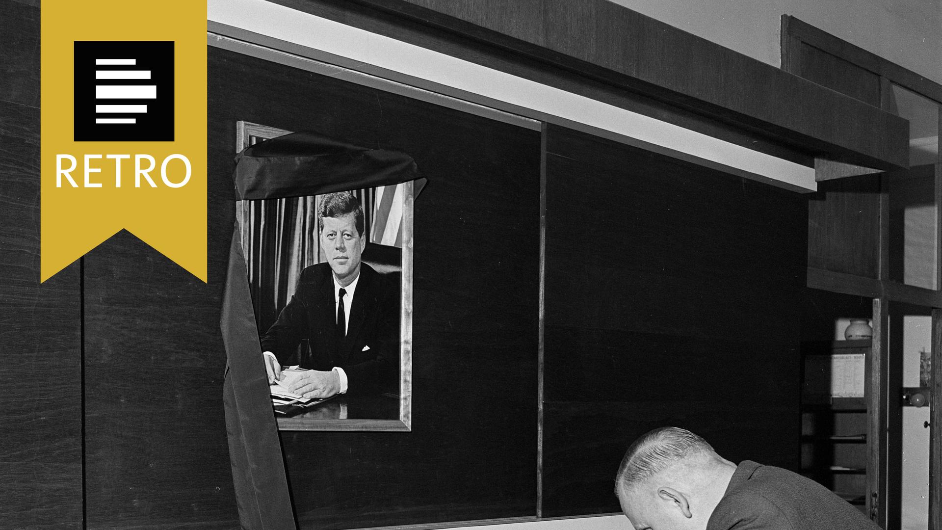 Ein Mann trägt sich in ein Kondulenzbuch ein. Im Hintergrund ein Bild von John F. Kennedy.