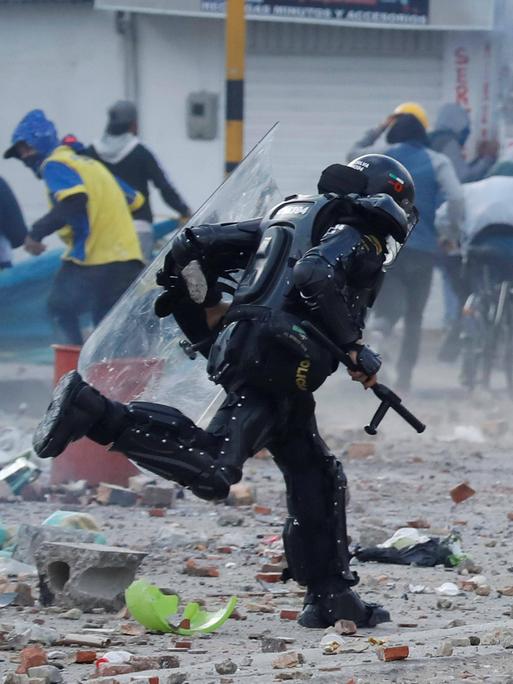 Ein Polizist in voller Schutzausrüstung und mit Schlagstock rennt auf einen Demonstranten zu. Im Hintergrund laufen viele Demonstranten weg.