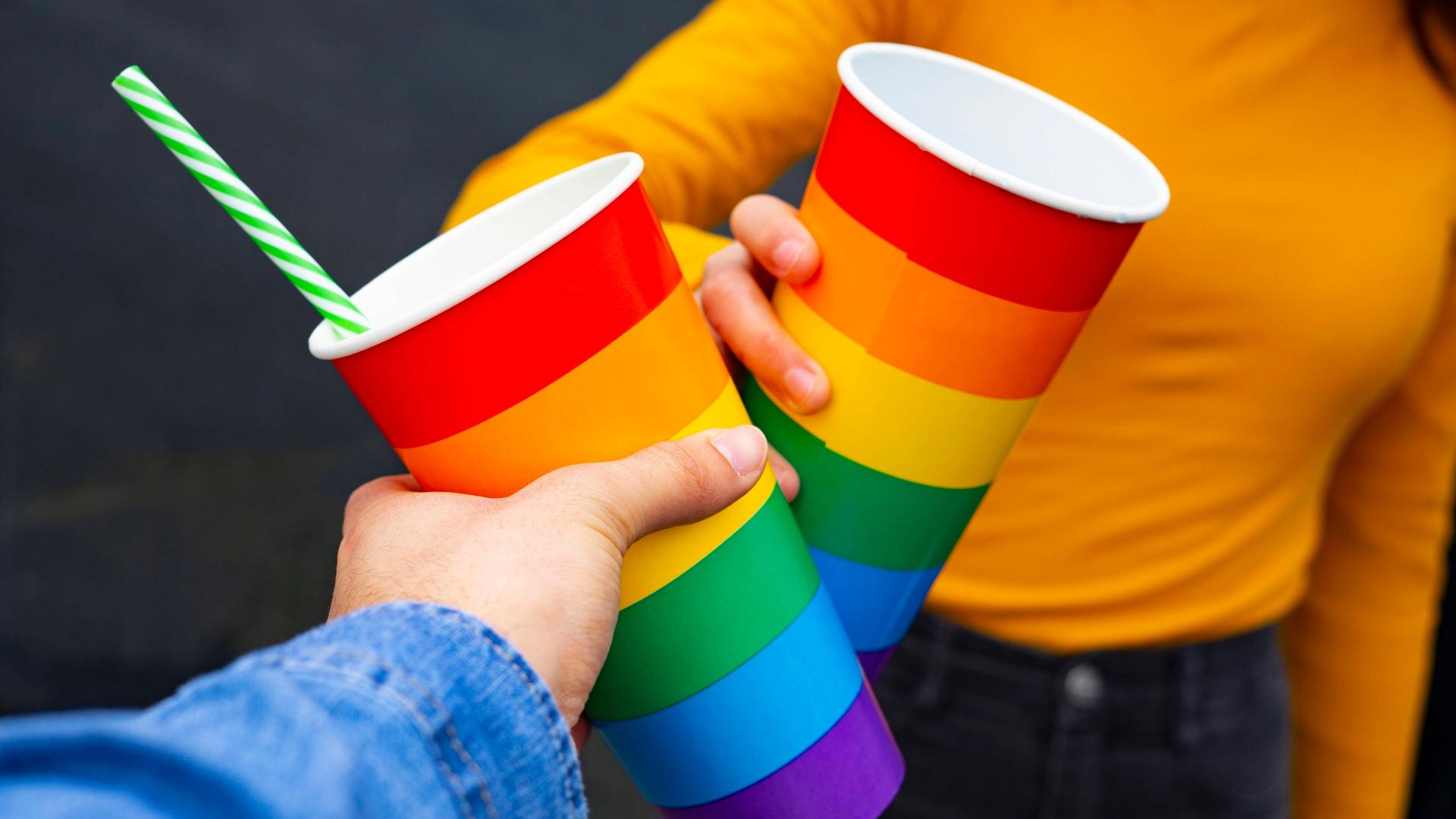 Zwei Menschen stoßen an mit Bechern in Regenbogenfarben. Die Farbkombination steht auch für die Vielfalt menschlichen Lebens und Liebens.