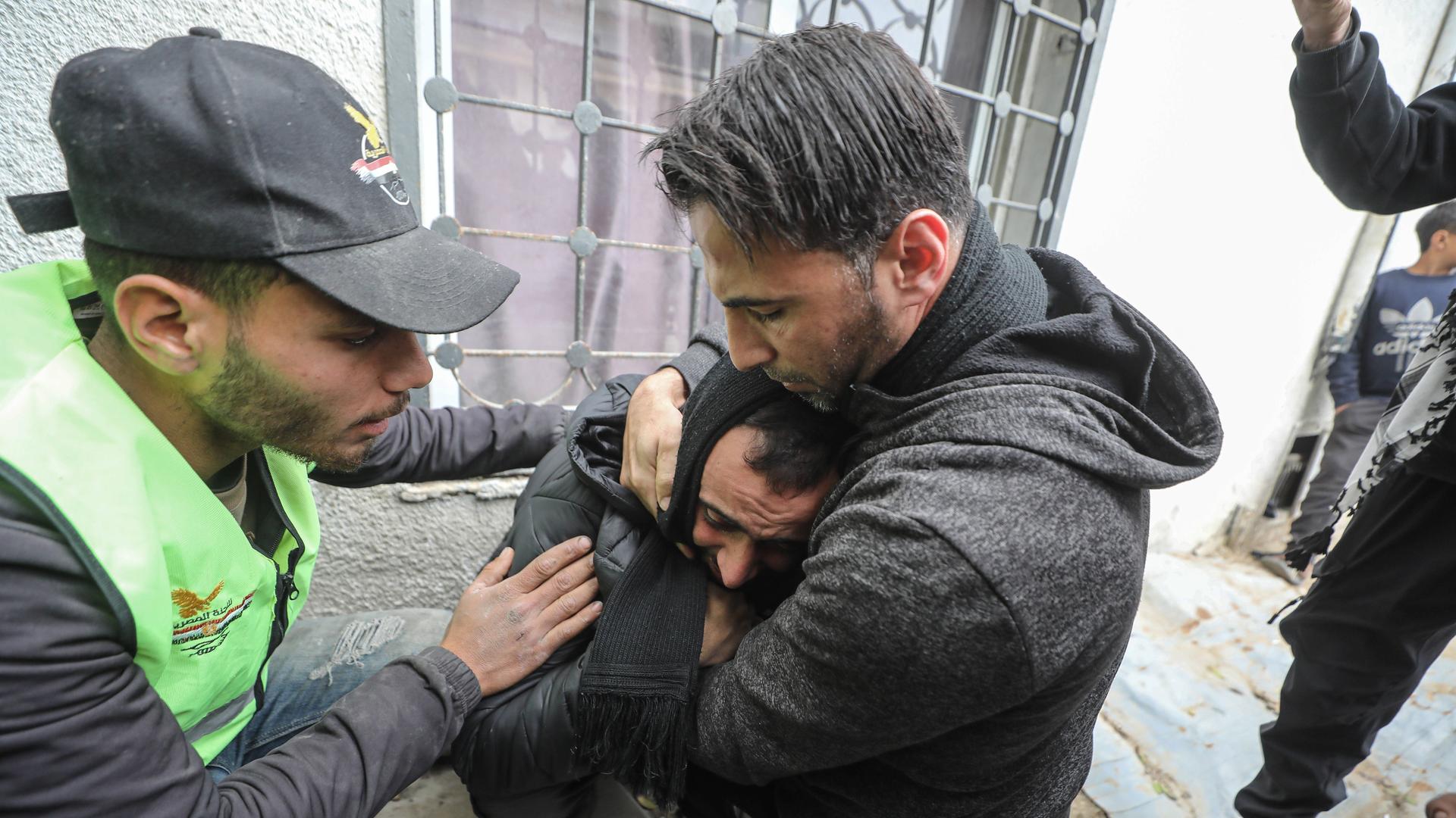 Ein trauriger Mann wird von einem anderen in den Arm genommen und getröstet. Ein anderer legt seine Hände um seine Schultern. 