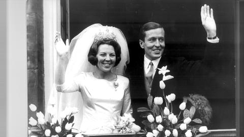 Prinzessin Beatrix von den Niederlanden heiratet den Baron Claus von Amsberg, dann Prinz Claus. Die Braut, links im Foto, träge ein weißes Kleid, der Bräutigam ein dunkles Sakko und Krawatte. Beide winken mit einer Hand.
