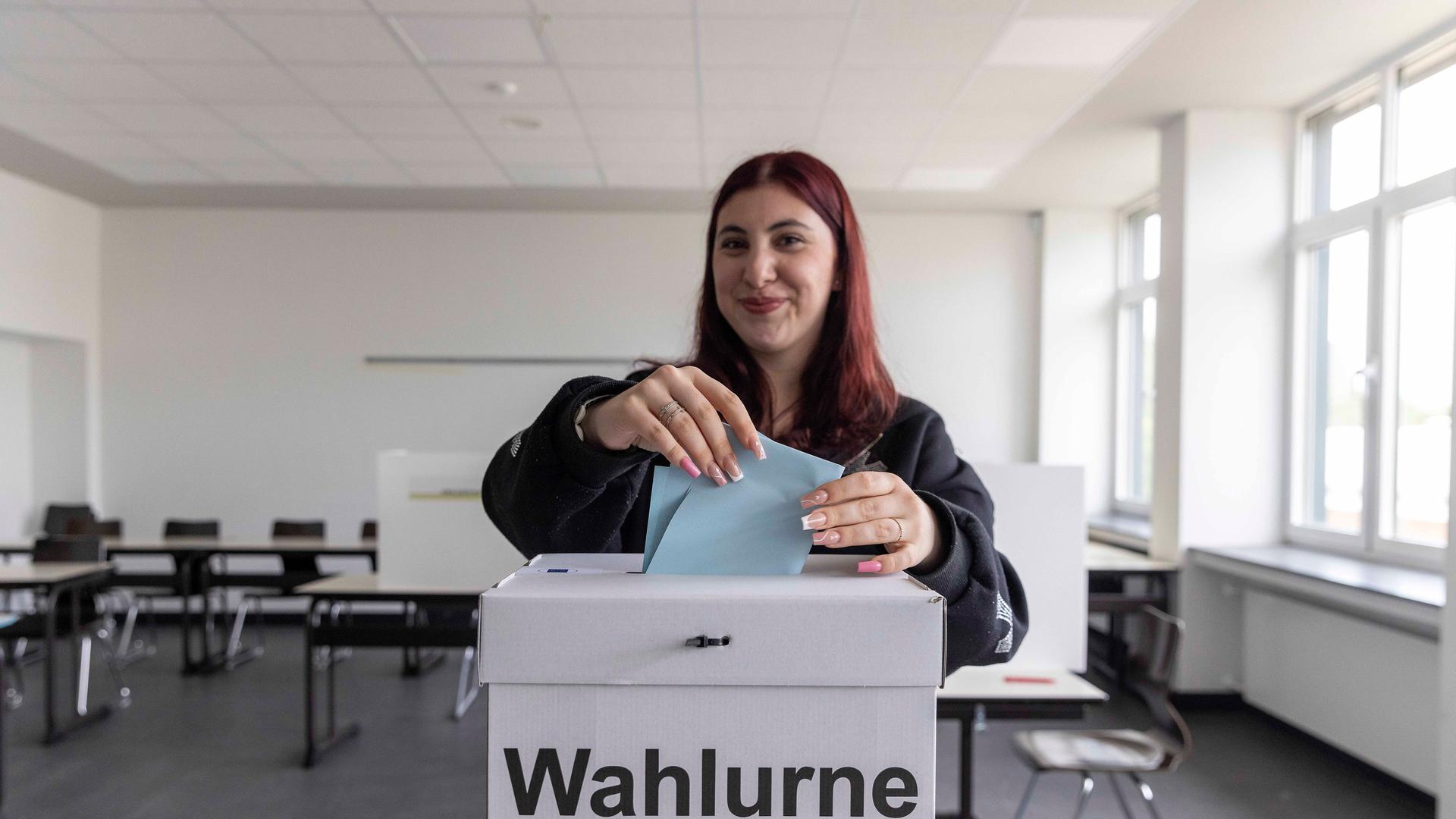 Eine junge Wählerin steht in einem Wahl-Raum und wirft einen Wahl-Zettel in eine Wahl-Urne.