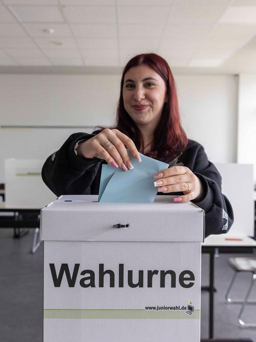 Eine junge Wählerin steht in einem Wahlraum und wirft einen Wahlzettel in eine Wahlurne.