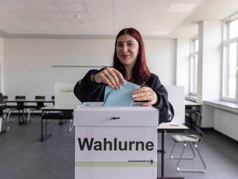 Eine junge Wählerin steht in einem Wahlraum und wirft einen Wahlzettel in eine Wahlurne.