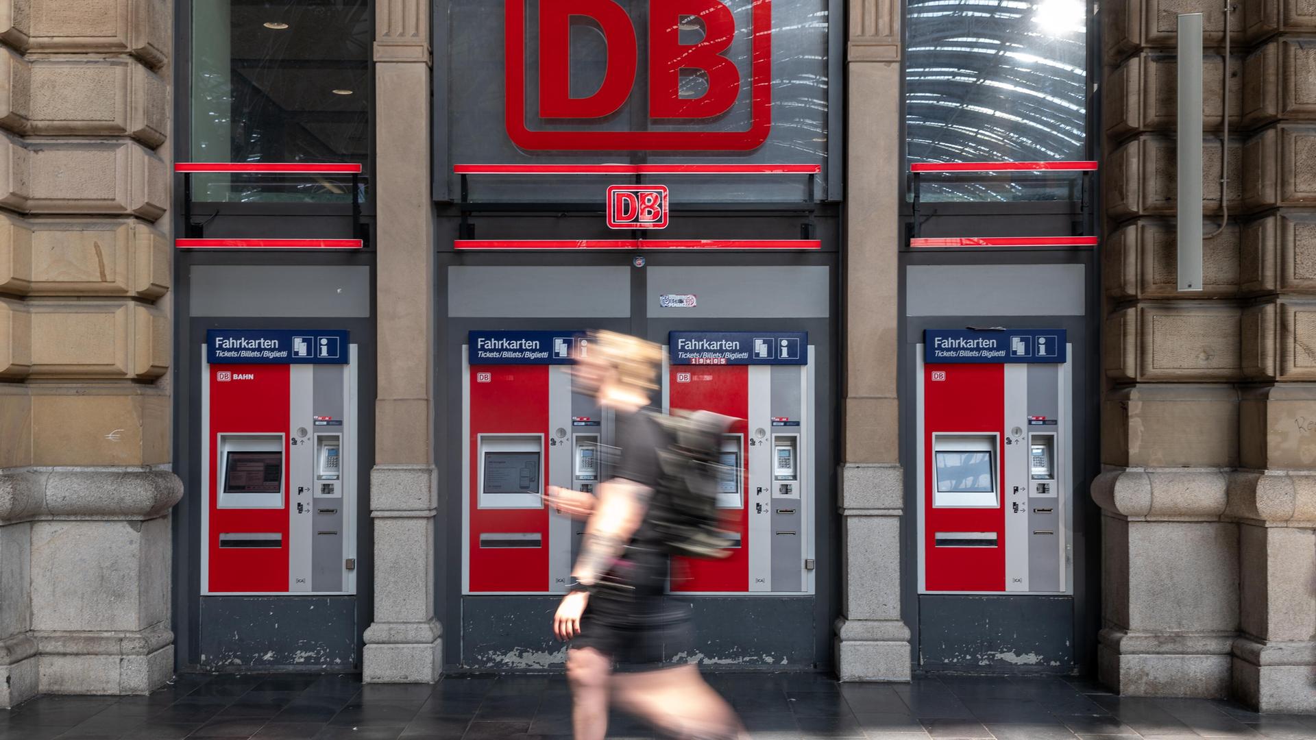 Vier rot-graue Fahrkartenautomaten der Deutschen Bahn stehen in einer Reihe unter einem großen, leuchtend roten DB-Logo in einer Bahnhofshalle.