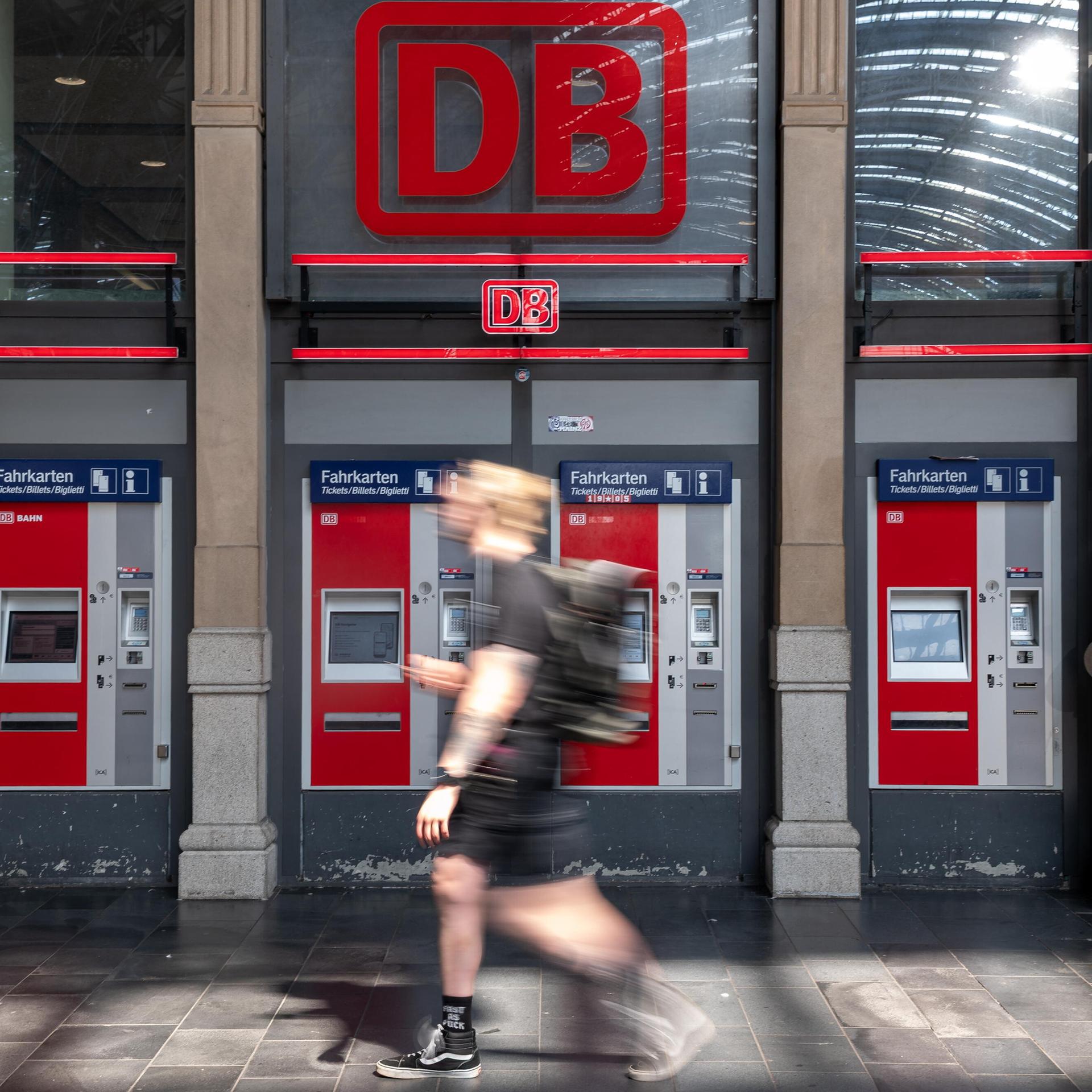 Vier rot-graue Fahrkartenautomaten der Deutschen Bahn stehen in einer Reihe unter einem großen, leuchtend roten DB-Logo in einer Bahnhofshalle.