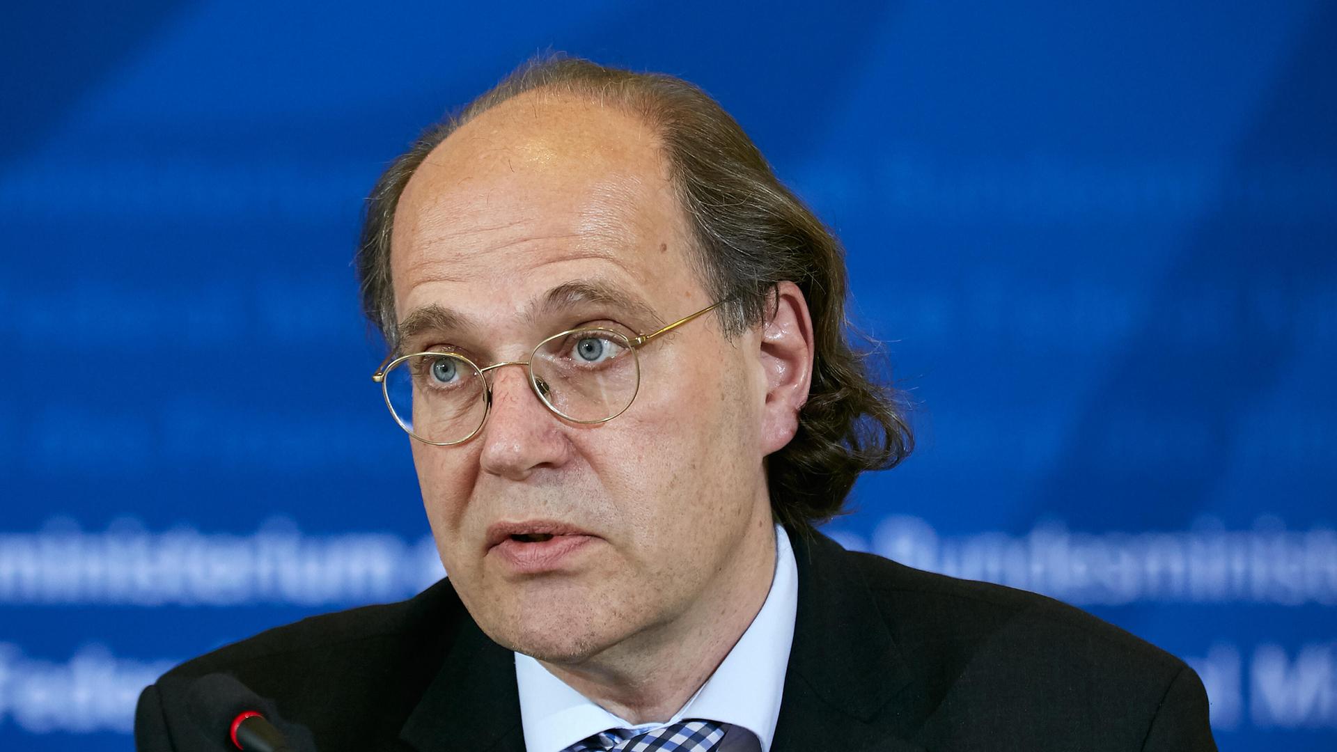 Das Bild zeigt den Wirtschaftswissenschaftler Thiess Büttner auf einer Konferenz. Er trägt einen schwarzen Anzug und ein blaues Hemd und schaut skeptisch in die Kamera.