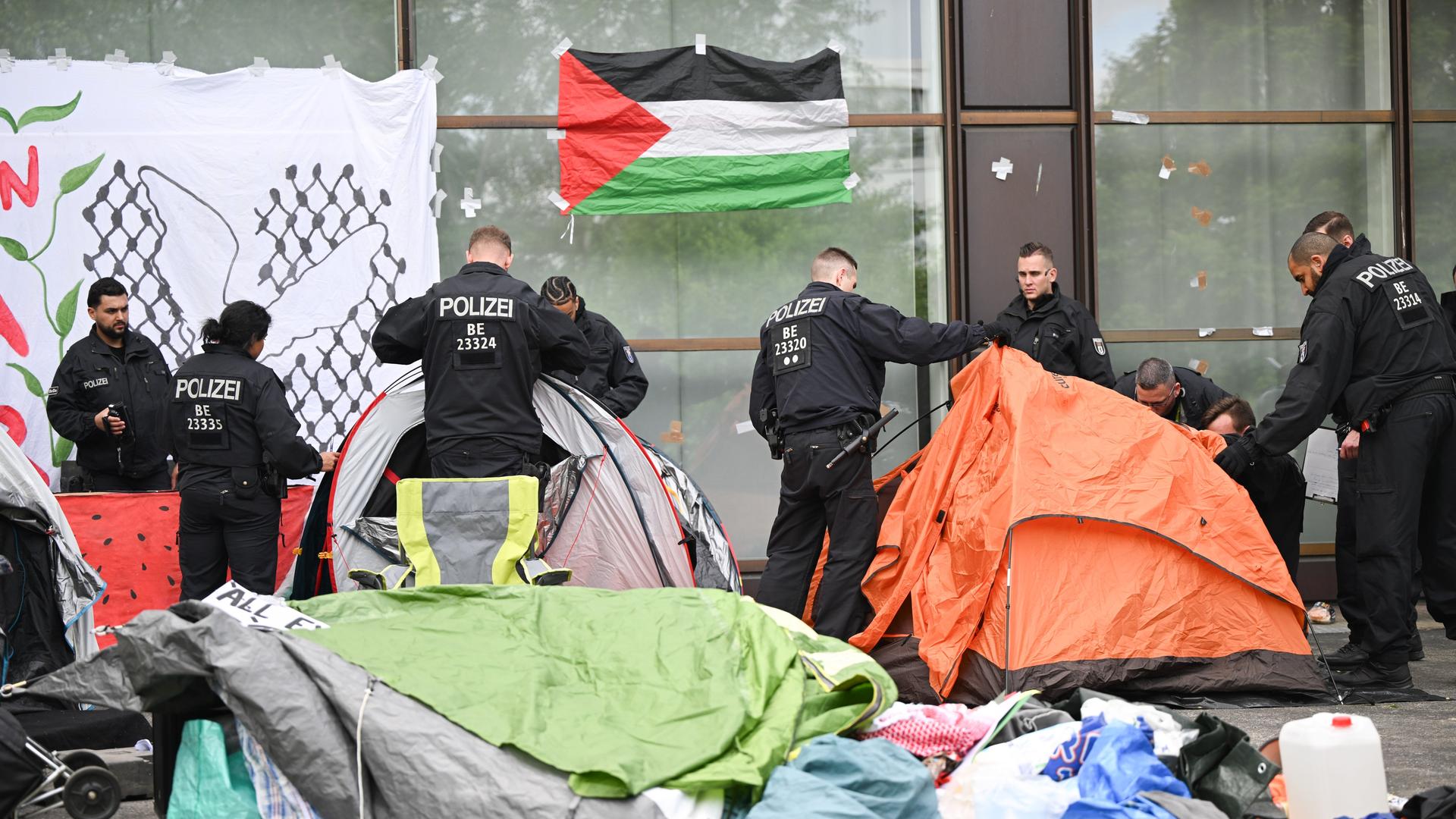 Polizisten räumen Zelte eines Protestcamps beiseite. An einer Wand hängt eine Palästina-Fahne.