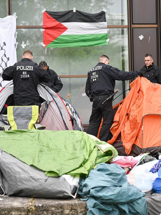Polizisten räumen Zelte eines Protestcamps beiseite. An einer Wand hängt eine Palästina-Fahne.