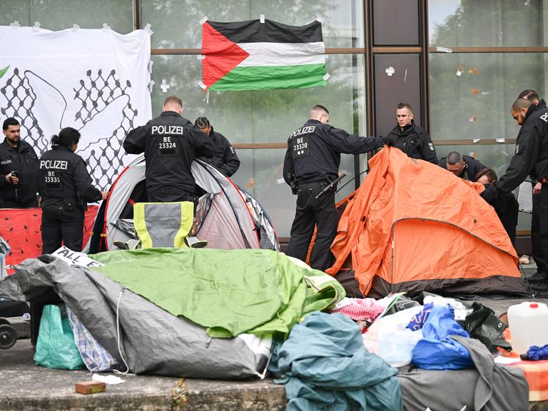 Polizisten räumen Zelte eines Protestcamps beiseite. An einer Wand hängt eine Palästina-Fahne.