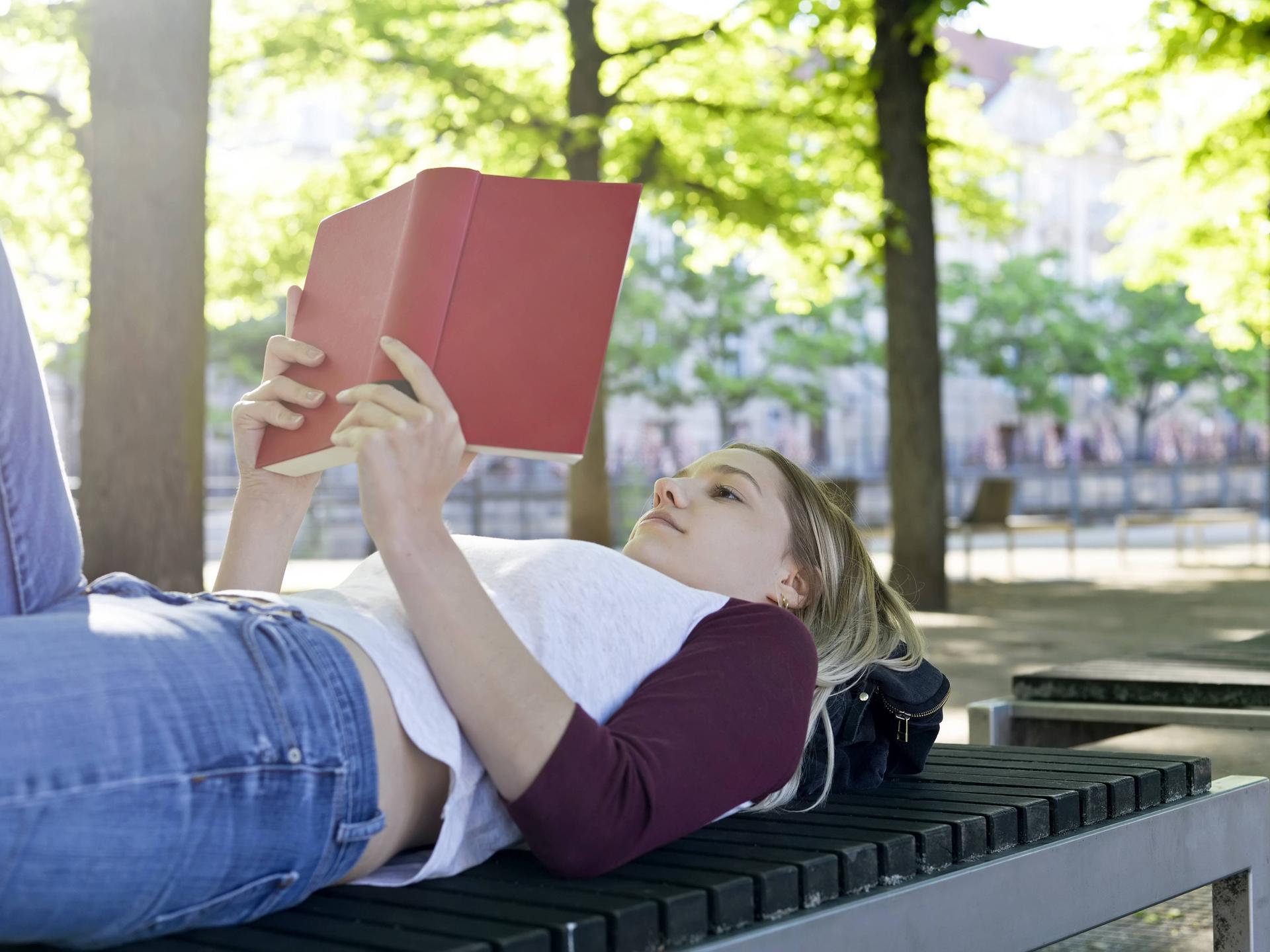Eine junge Frau liegt auf eine Parkbank und liest entspannt ein Buch (Symbolfoto) Eine junge Frau liegt auf eine Parkbank und liest entspannt ein Buch (Symbolfoto)