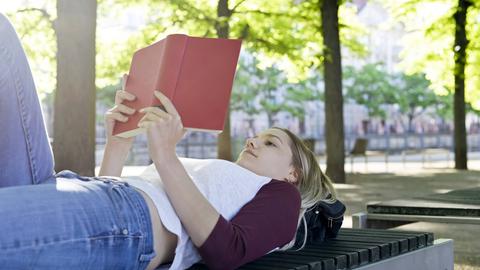 Eine junge Frau liegt auf eine Parkbank und liest entspannt ein Buch (Symbolfoto) Eine junge Frau liegt auf eine Parkbank und liest entspannt ein Buch (Symbolfoto)