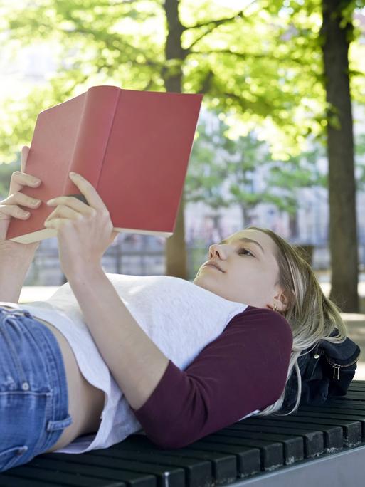 Eine junge Frau liegt auf eine Parkbank und liest entspannt ein Buch (Symbolfoto) Eine junge Frau liegt auf eine Parkbank und liest entspannt ein Buch (Symbolfoto)