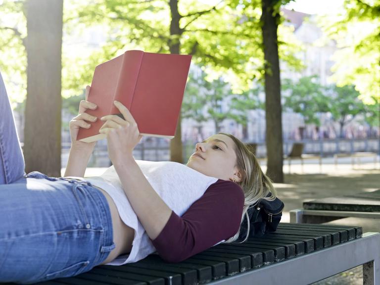 Eine junge Frau liegt auf eine Parkbank und liest entspannt ein Buch (Symbolfoto) Eine junge Frau liegt auf eine Parkbank und liest entspannt ein Buch (Symbolfoto)
