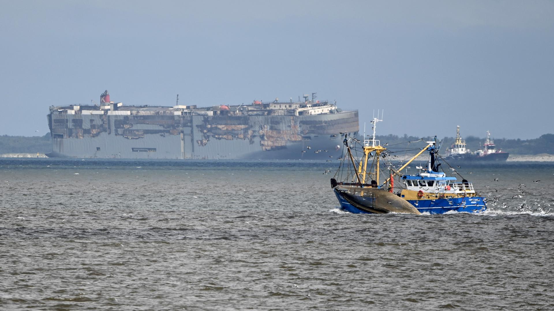 Das Bild zeigt den Frachter und den Schlepper in der Ferne unter graublauem Himmel vor der Küste. Im Hintergrund ein Fischerboot. Das Bild zeigt den Frachter und den Schlepper in der Ferne unter graublauem Himmel vor der Küste. Im Hintergrund ein Fischerboot.