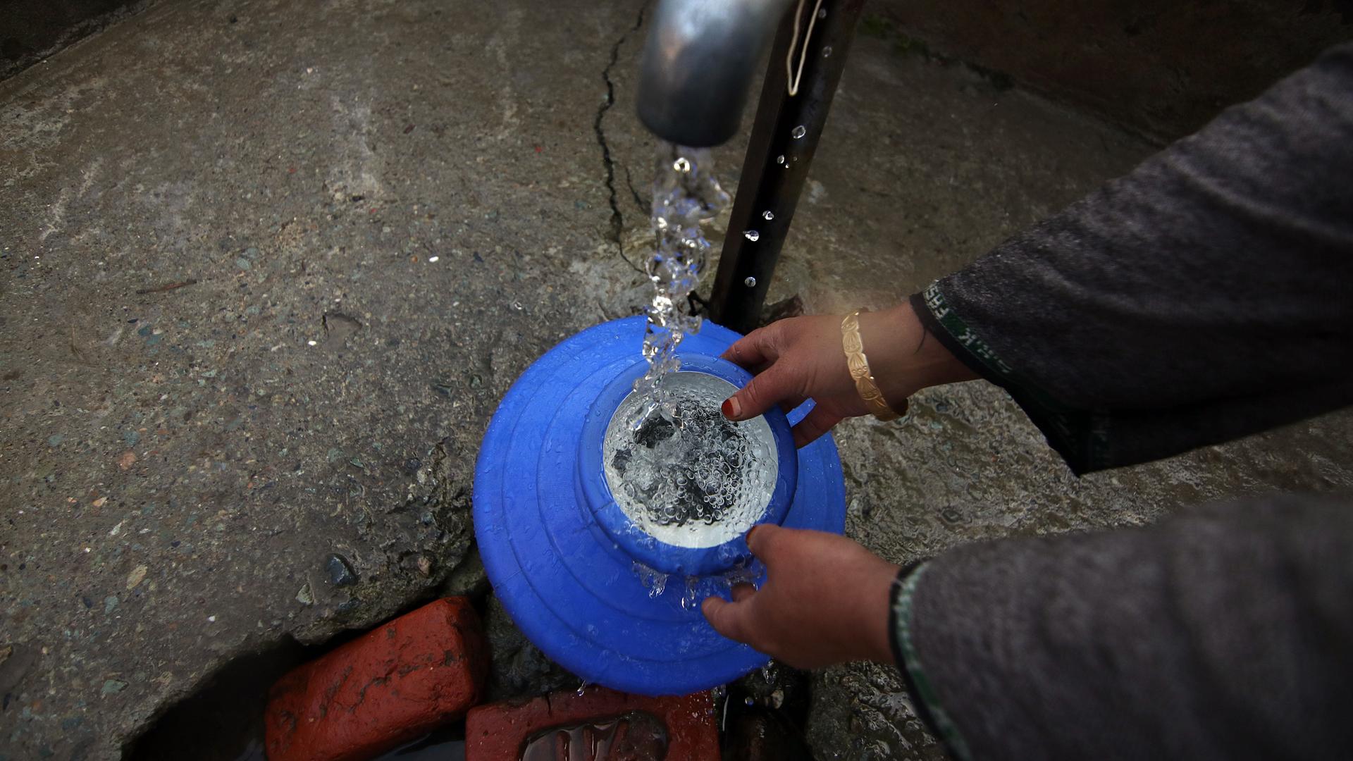 Eine Frau hält einen blauen Topf unter einen sprudelnden Wasserhahn. Das Wasser füllt den Topf.