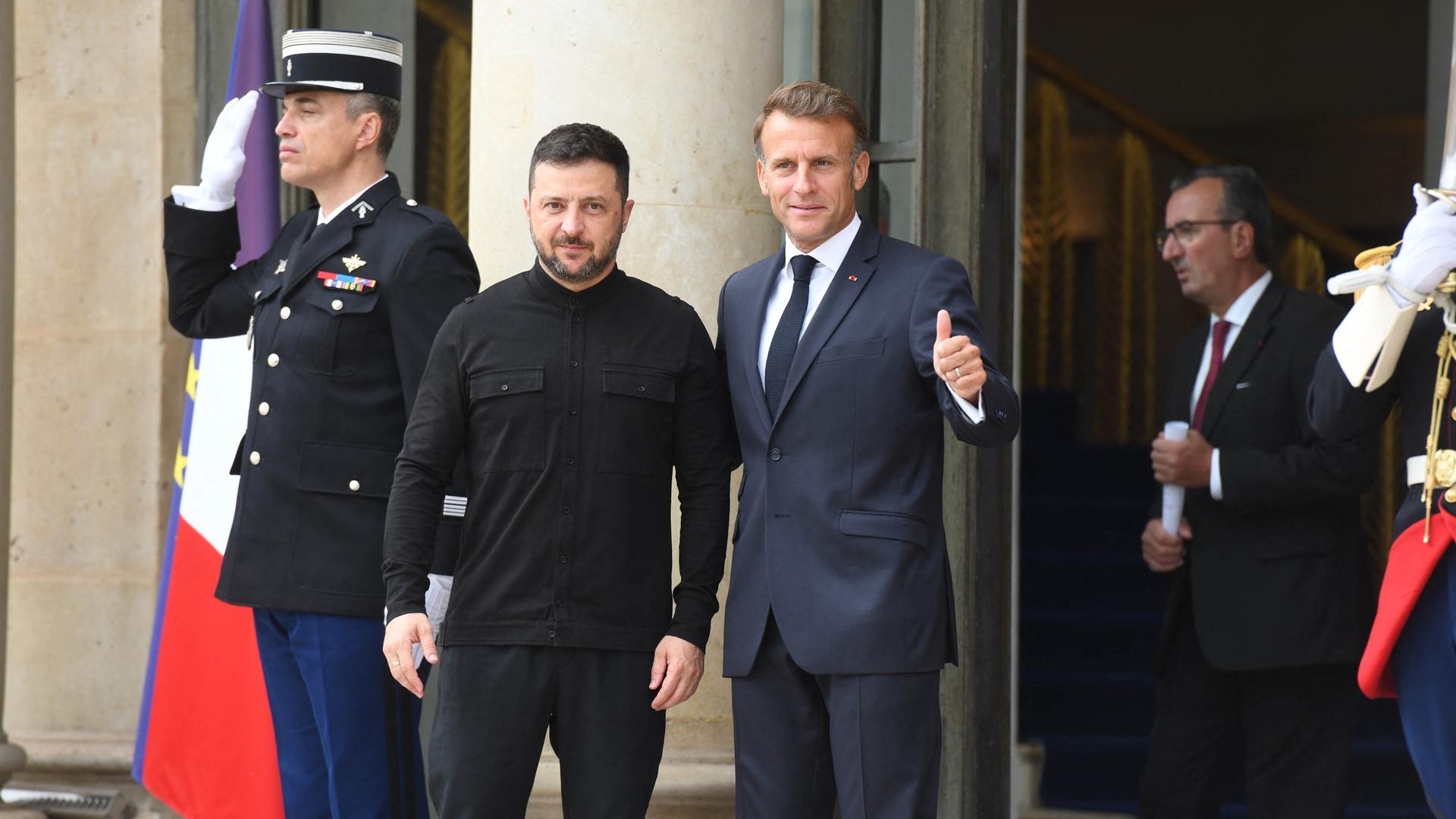 Frankreichs Präsident Macron hält den ukrainische Präsident Selenskyj am Arm und hebt mit der anderen Hand den Daumen, beide lächeln.