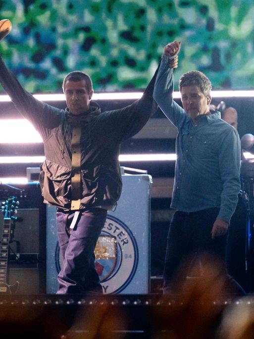 Liam Gallagher (l.), hält die Hand von Noel Gallagher von der Band Oasis während ihres Reunion-Konzerts am Freitag, 4. Juli 2025, in Cardiff