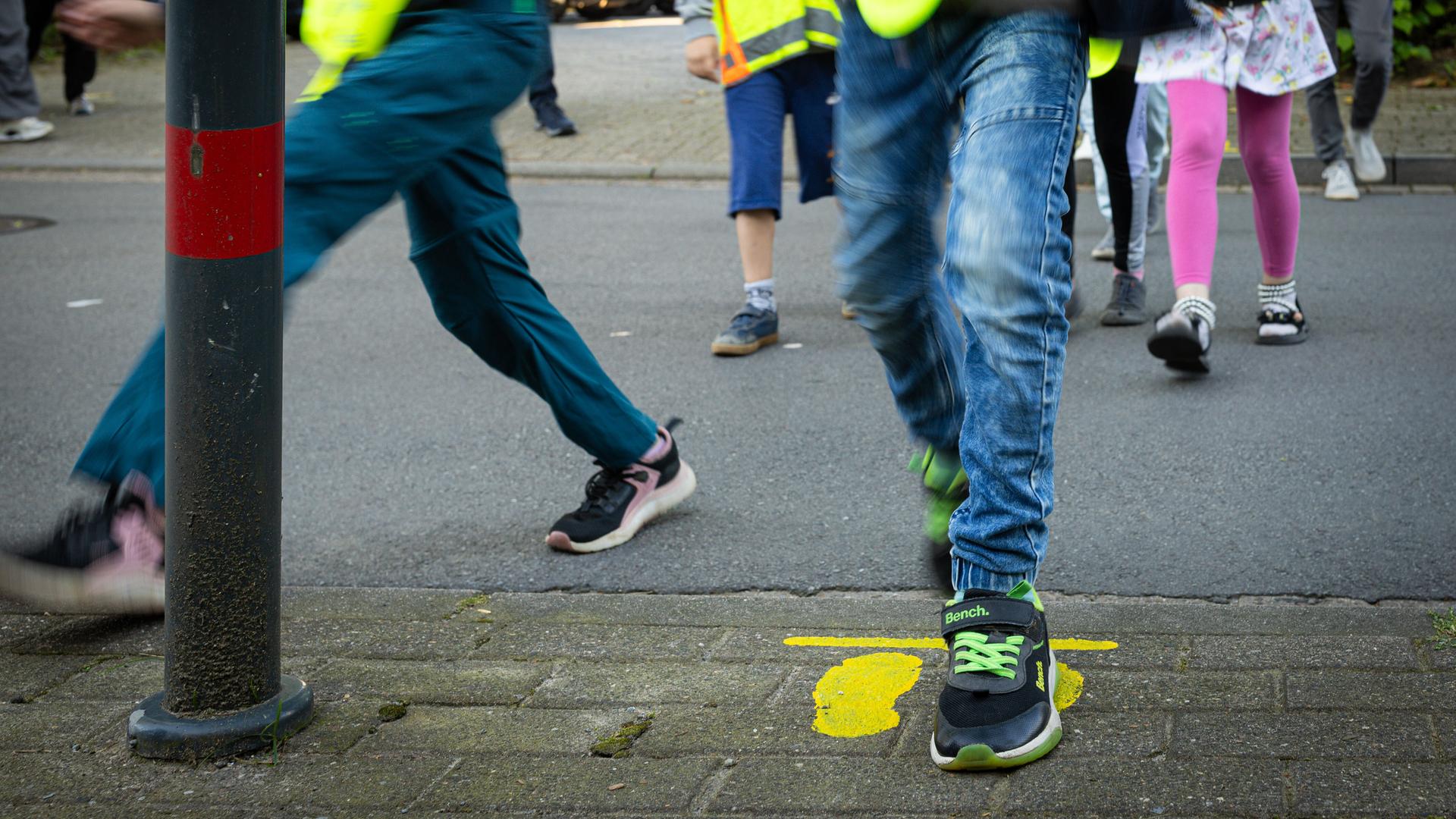 Auf einem Gehweg an einer Verkehrsstraße sind gelbe Füße angezeichnet. Kinder laufen über die Straße.