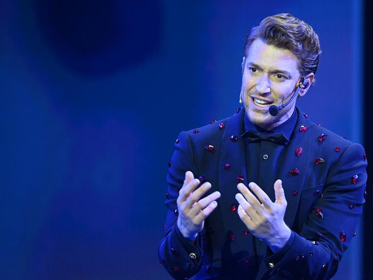 Daniel Donskoy steht im Anzug auf der Bühne und spricht in ein Mikrofon.