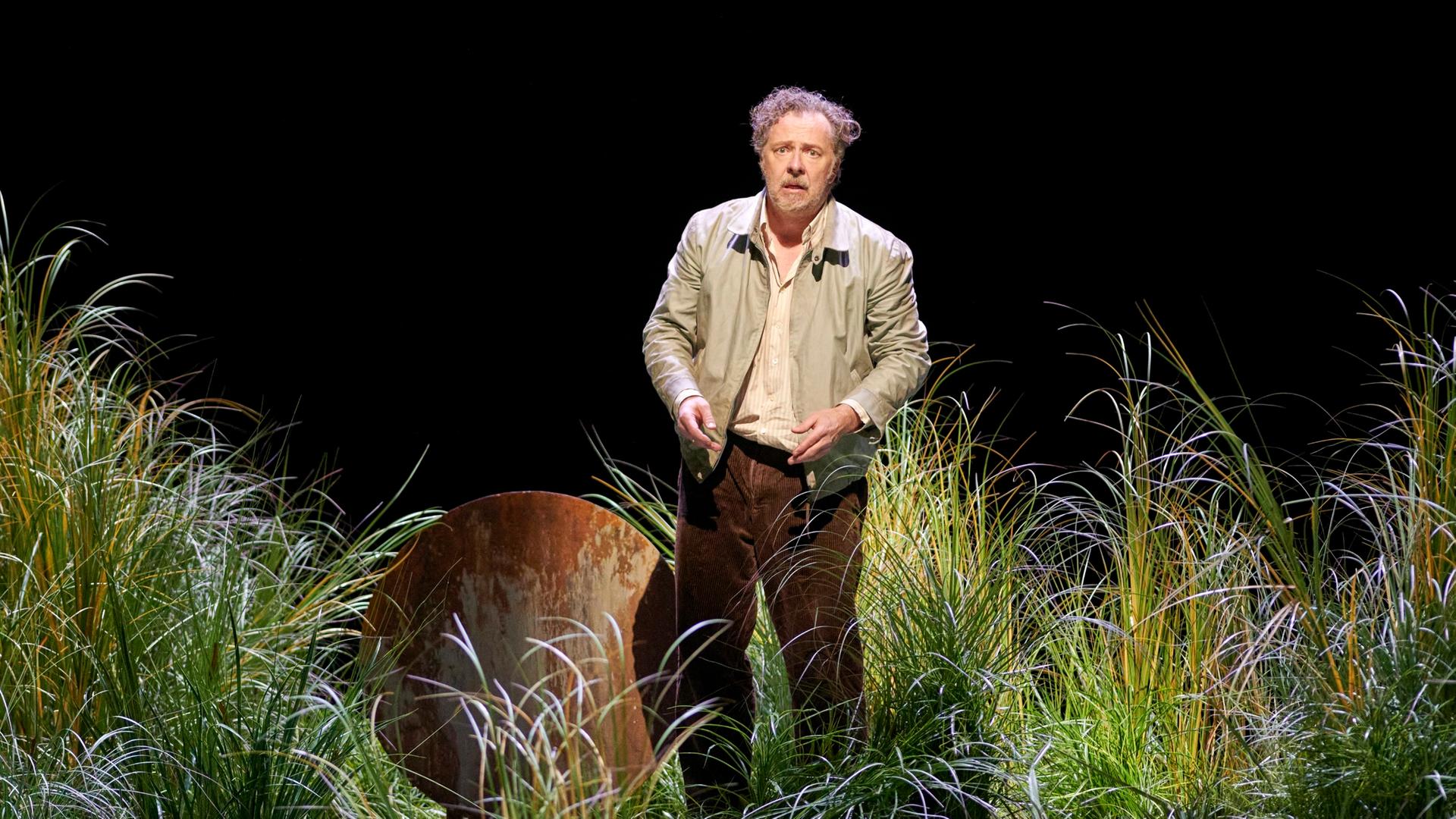 Ein Mann mit grauem, zerzaustem Haar steht auf einer Bühne. Er ist umgeben von hohem Gras und guck verwirrt. Er träge eine beige Jacke. 