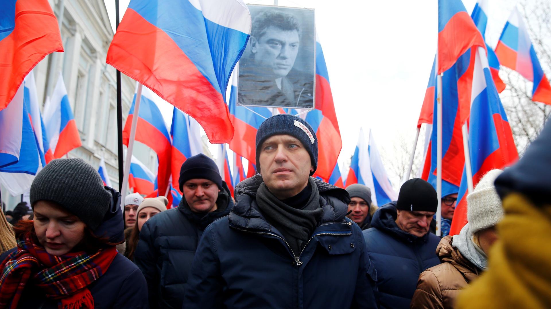 Alexej Nawalny bei einem Marsch in Gedenken an den oppositionellen Boris Nemtsow 2015. Er trägt eine dunkle Wollmütze und eine dunkelblaue Winterjacke. Hinter ihm gehen viele Menschen, die zum Teil russische Fahnen hochhalten. Jemand hält ein an einem Stab befestigte Schwarz-Weiß-Foto von Boris Nemtsow hoch.