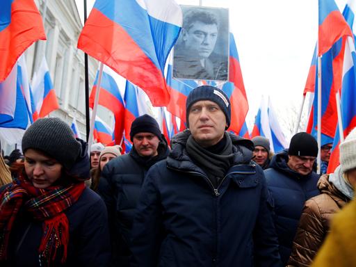 Alexej Nawalny bei einem Marsch in Gedenken an den oppositionellen Boris Nemtsow 2015. Er trägt eine dunkle Wollmütze und eine dunkelblaue Winterjacke. Hinter ihm gehen viele Menschen, die zum Teil russische Fahnen hochhalten. Jemand hält ein an einem Stab befestigte Schwarz-Weiß-Foto von Boris Nemtsow hoch.