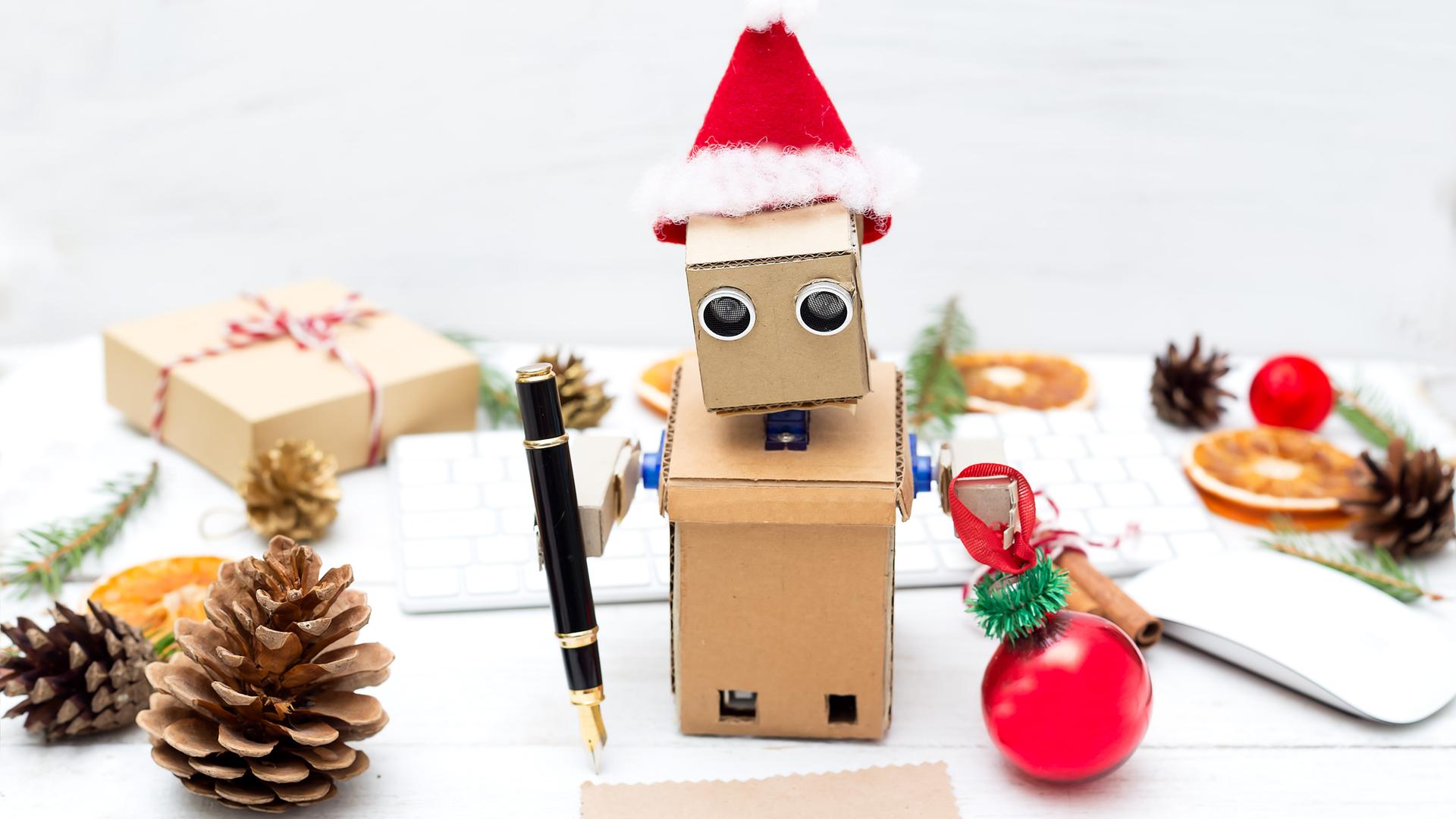 Ein aus Pappe gebastelter Roboter mit Weihnachtsmannmütze schreibt mit einem Füller auf ein Stückchen Papier. 