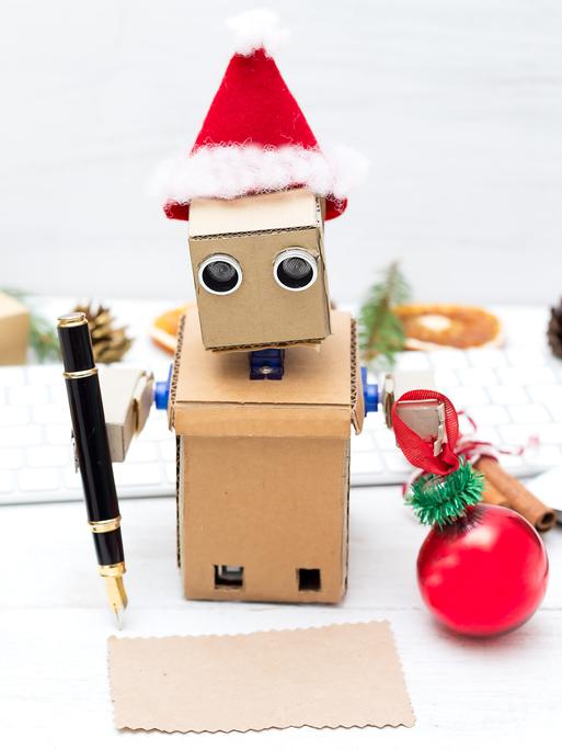 Ein aus Pappe gebastelter Roboter mit Weihnachtsmannmütze schreibt mit einem Füller auf ein Stückchen Papier. 