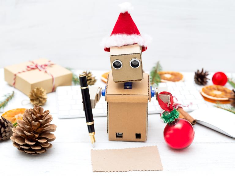 Ein aus Pappe gebastelter Roboter mit Weihnachtsmannmütze schreibt mit einem Füller auf ein Stückchen Papier. 