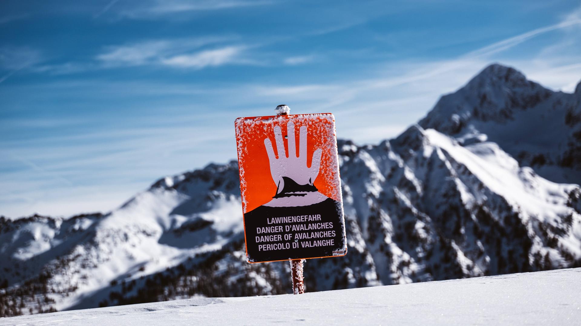 Ein rotes Schild mit einer erhobenen Hand warnt Skifahrer vor einer Bergkulisse vor der Lawinengefahr.