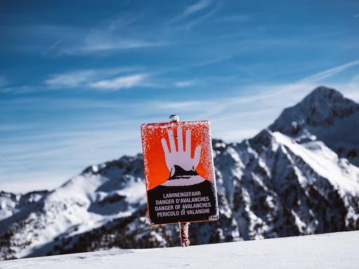 Ein rotes Schild mit einer erhobenen Hand warnt Skifahrer vor einer Bergkulisse vor der Lawinengefahr.
