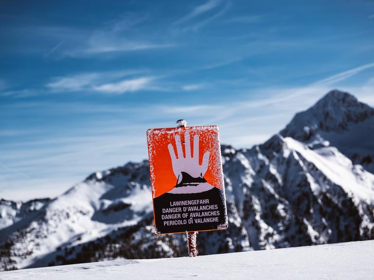 Ein rotes Schild mit einer erhobenen Hand warnt Skifahrer vor einer Bergkulisse vor der Lawinengefahr.