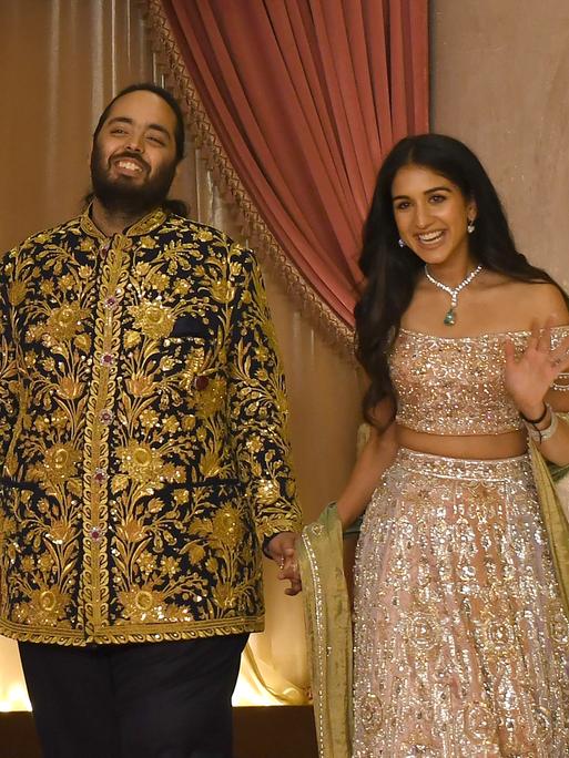Der indische Milliardärssohn Anant Ambani und seine Verlobte Radhika Merchant stehend lächelnd nebeneinander, sie winkt in die Kamera. 