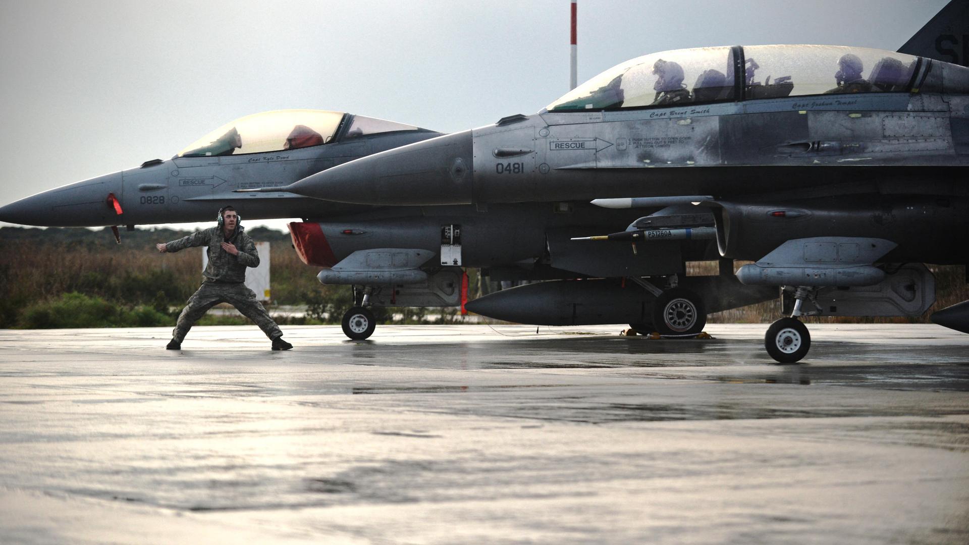 Zwei Kampflieger F-16 Fighting Falcon auf einem Flugplatz, ein Soldat steht breitbeinig vor einem der Flugzeuge und gibt ein Kommando
