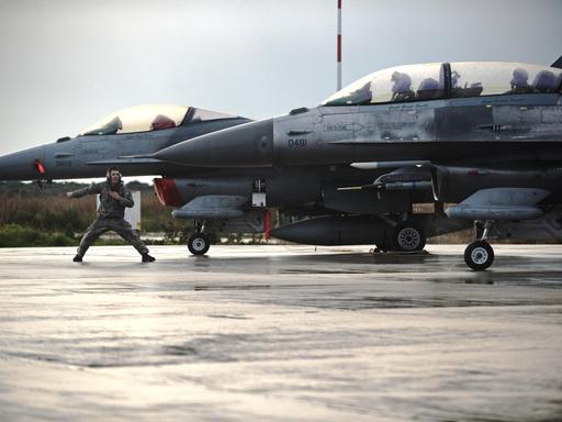 Zwei Kampflieger F-16 Fighting Falcon auf einem Flugplatz, ein Soldat steht breitbeinig vor einem der Flugzeuge und gibt ein Kommando