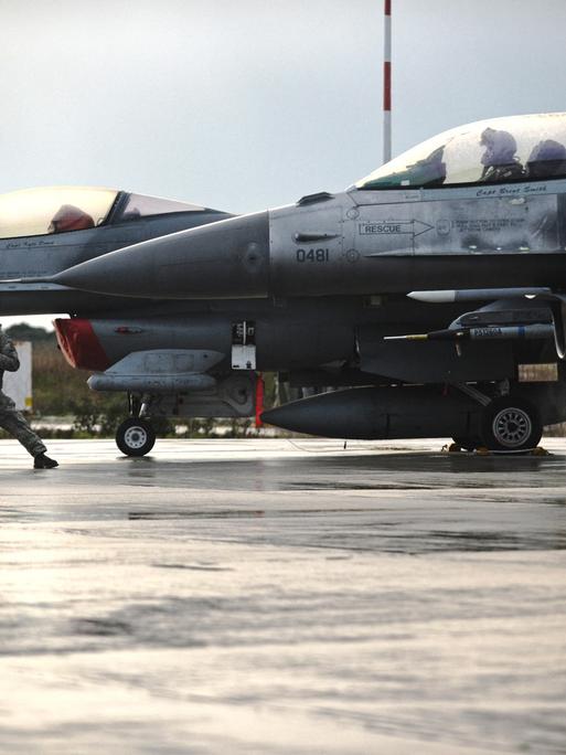 Zwei Kampflieger F-16 Fighting Falcon auf einem Flugplatz, ein Soldat steht breitbeinig vor einem der Flugzeuge und gibt ein Kommando
