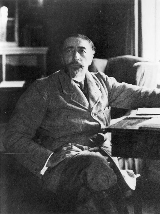 Der Schriftsteller Joseph Conrad (1857-1924) sitzt mit übergeschlagenen Beinen seitlich an einem Schreibtisch.