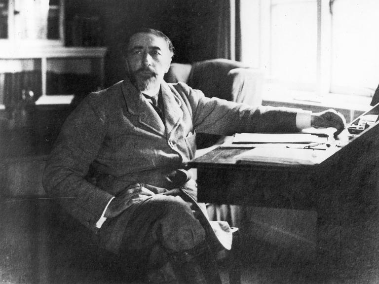 Der Schriftsteller Joseph Conrad (1857-1924) sitzt mit übergeschlagenen Beinen seitlich an einem Schreibtisch.