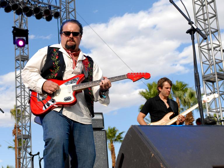 Ein Mann mit Weste und Sonnenbrille spielt auf einer Bühne Gitarre